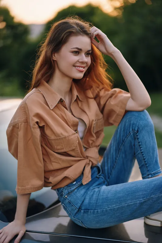 Young woman in casual attire sitting on car hood, smiling with sunset background, showcasing relaxed summer vibe and fashion style.