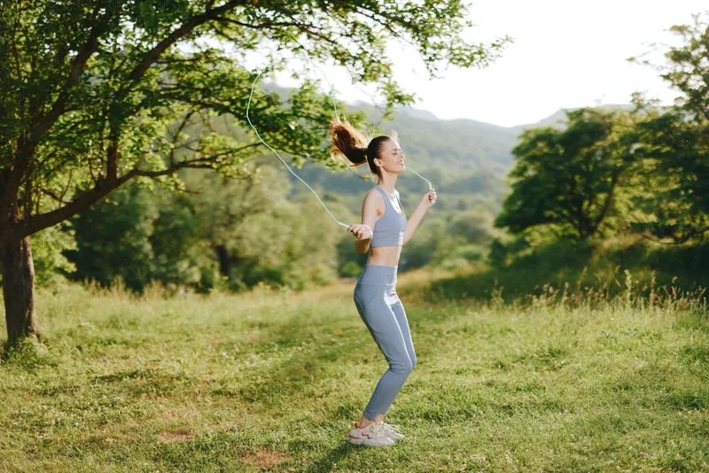 Young woman jumping rope outdoors in activewear, enjoying fitness in nature, vibrant green scenery, healthy lifestyle concept