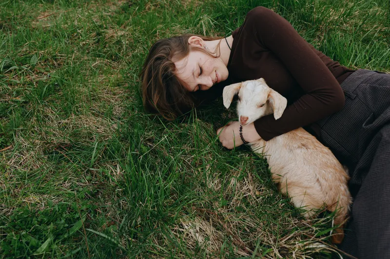 Young woman lying on grass gently hugs a baby goat showing tender care and connection in a natural outdoor lifestyle moment during a peaceful day.