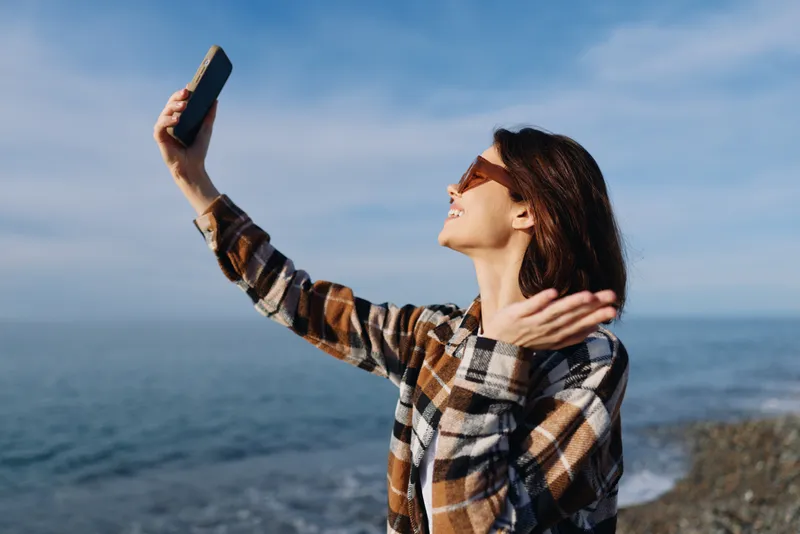 Woman taking a selfie by the sea in a plaid shirt, sunny day, wind in hair, ocean horizon, casual outdoor portrait for lifestyle, travel and leisure visuals
