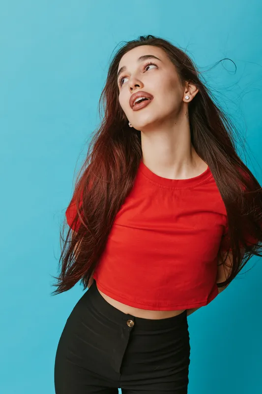 Young woman with long hair wearing red crop top and black pants posing against blue background with expressive face and casual style for fashion and lifestyle concept.