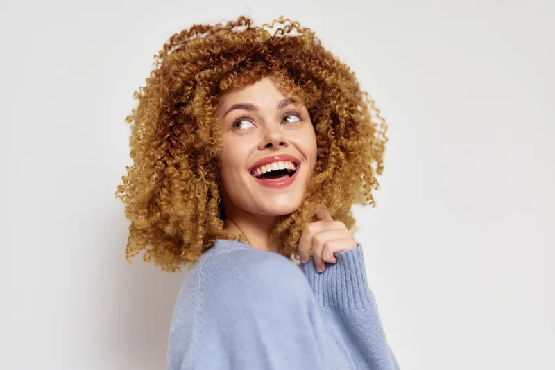 Счастливая женщина с вьющимися светлыми волосами улыбается в уютном вязаном свитере