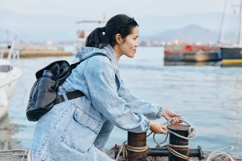Harbor setting with a focused woman in denim jacket adjusting mooring ropes, preparing boat docking, outdoor waterfront scene, casual seafront work and travel lifestyle