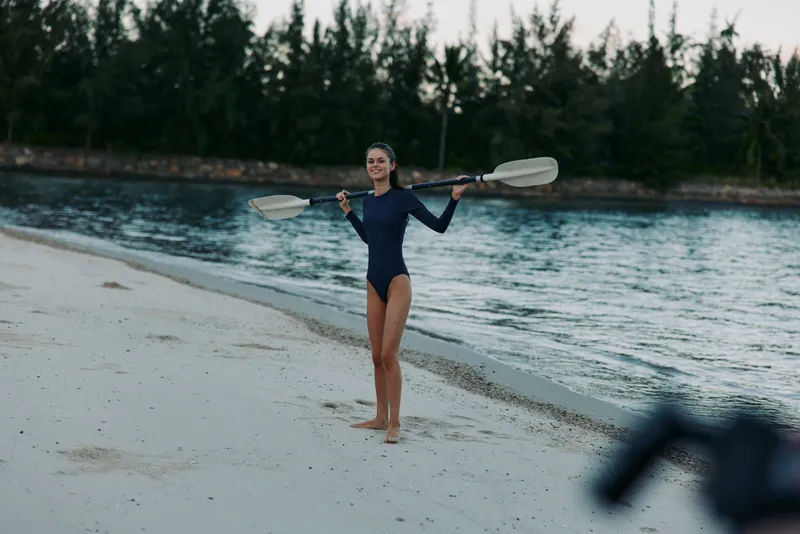 Female athlete stands on sandy beach wearing a swimsuit, balancing a paddle across shoulders, ready for water sport action by tranquil sea and forested shoreline during early light, confident pose.