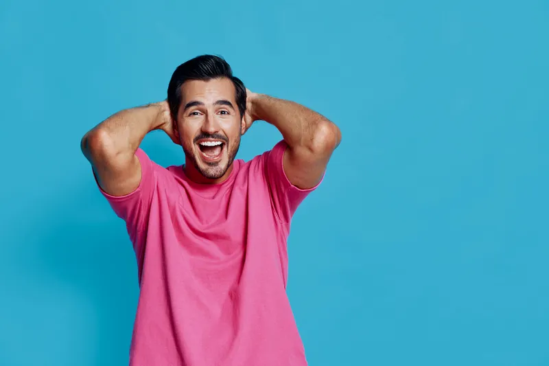 Full body joyful smiling young man wearing pink t-shirt posing with hands behind head on solid blue background, capturing a fun and lively moment.