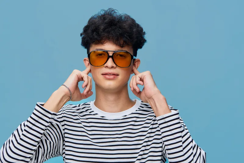 Young man wearing striped shirt and orange sunglasses covering ears with fingers against blue background, expressing playful attitude and casual style.