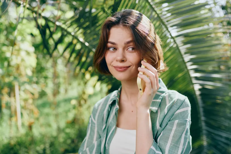Young woman outdoors, wearing a light green striped shirt over a white top, smiling softly while touching her ear.
