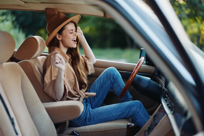 Happy young woman singing while driving in a vintage car, wearing a stylish hat, enjoying the moment in a sunny outdoor setting with vibrant green foliage.
