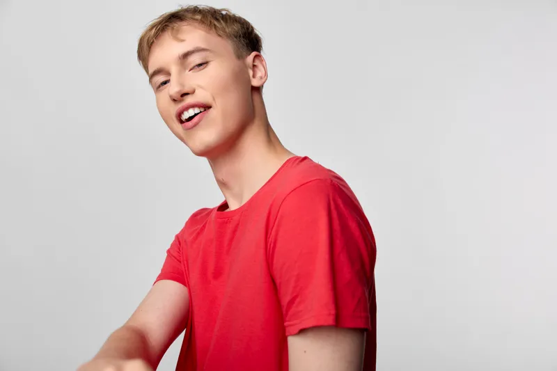 Young man in a red shirt with a bright smile performs a casual pose in a clean studio setting, radiating friendly expression and approachable energy for lifestyle or fashion projects