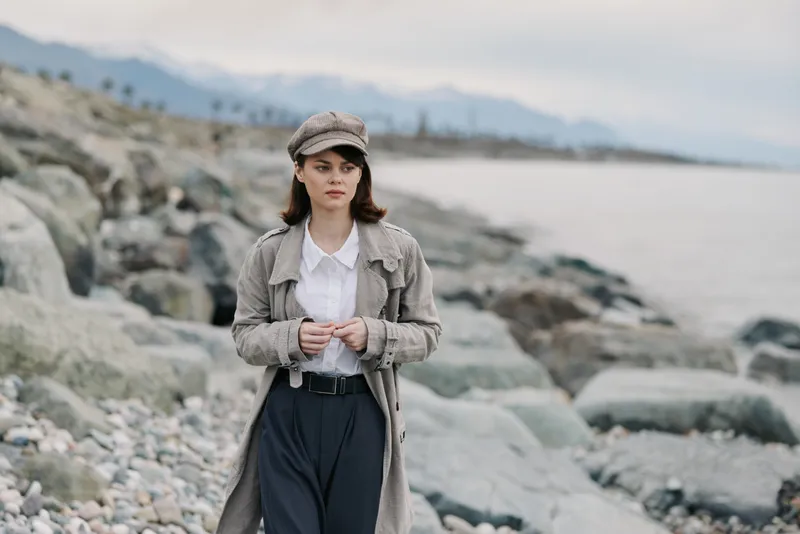 Young woman in vintage coat and cap walks on rocky beach near sea under cloudy sky. Casual autumn style and outdoor leisure concept with natural coastal landscape background.