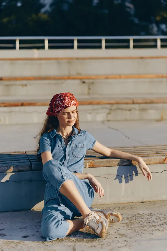 Lifestyle woman in modern boho-western denim street style wearing a red bandana, sitting outdoors in warm film color tones, relaxed and casual mood.