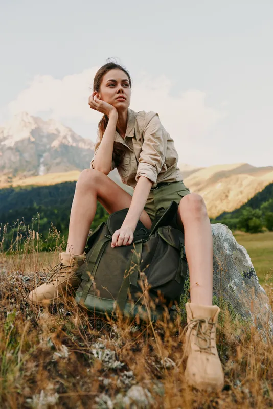Mountain hike scene with a young woman resting on a rock, wearing a green skirt and boots, a backpack nearby, gazing thoughtfully into the distance under soft daylight in wide open countryside