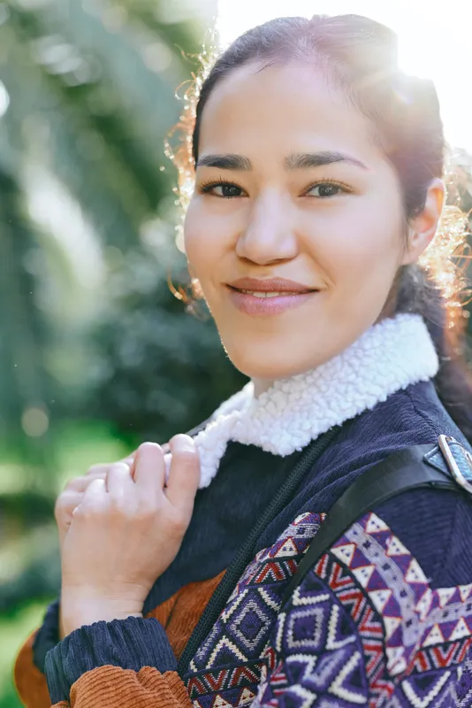 Smiling woman outdoors wearing a patterned sweater and jacket, natural light enhances greenery, casual fashion and lifestyle mood with warm colors
