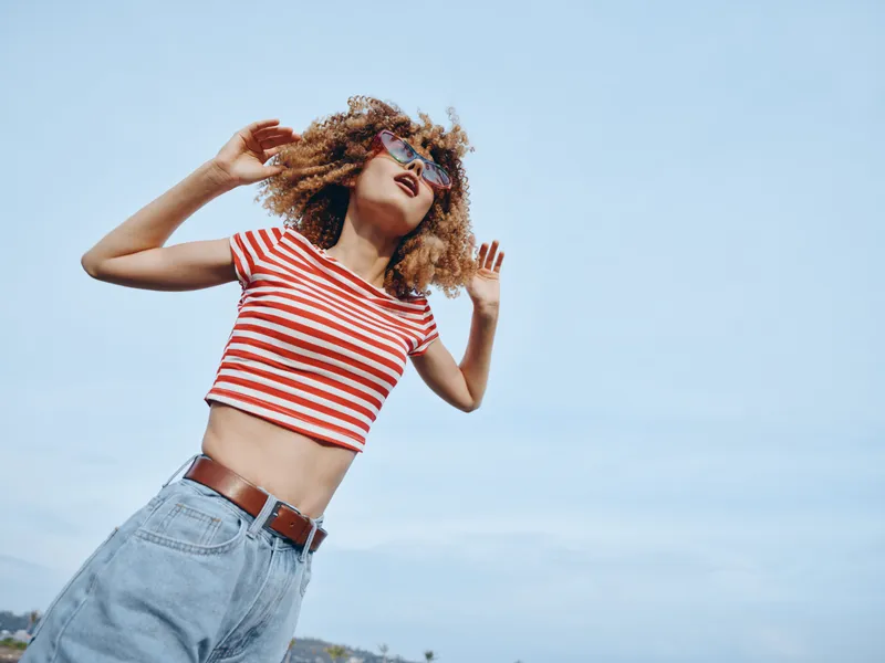 Carefree young woman with curly hair wearing a red and white striped crop top and denim jeans, standing against a bright blue sky, capturing a playful sunlit moment.