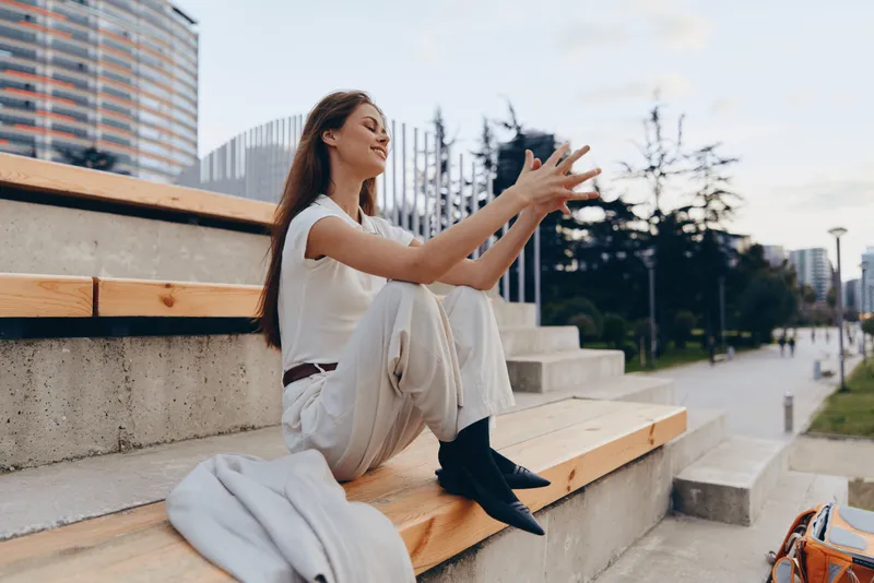 Young woman in casual outfit sitting on urban bench smiling with peaceful emotions, city background, autumn outdoor fashion style