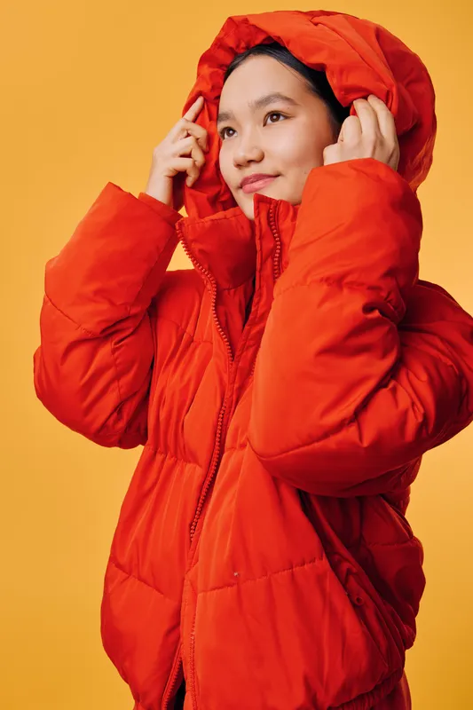 Woman wearing bright red warm jacket with hood, posing on orange background. Female model adjusting hood, smiling and dressed in winter outerwear with zipper and pockets.