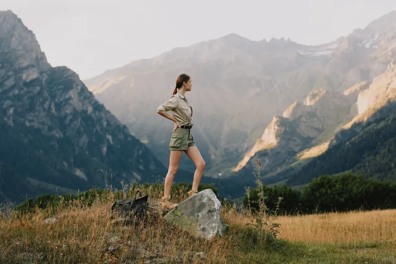 Young woman in hiking shorts and beige top stands on a rock amid a rugged mountain valley, enjoying a moment of exploration and freedom with a sense of adventure and calm as the wind sweeps the grass.