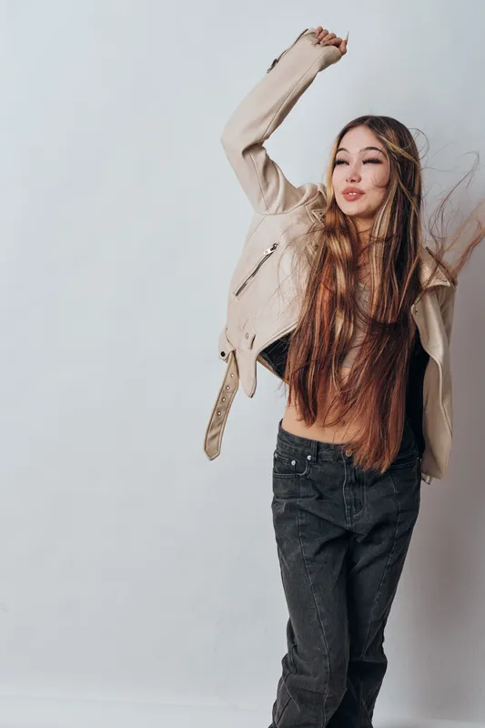 Young woman with long hair wearing beige jacket and black pants dancing with eyes closed and arms raised against white background in studio, casual style and joyful expression.