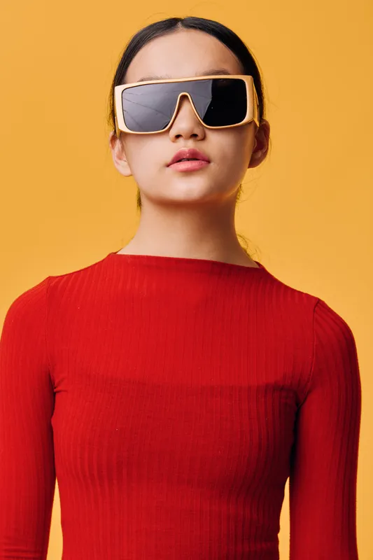Young woman wearing oversized sunglasses and a red ribbed sweater standing against yellow background. Fashion style, confident pose with minimal makeup and casual hairstyle.