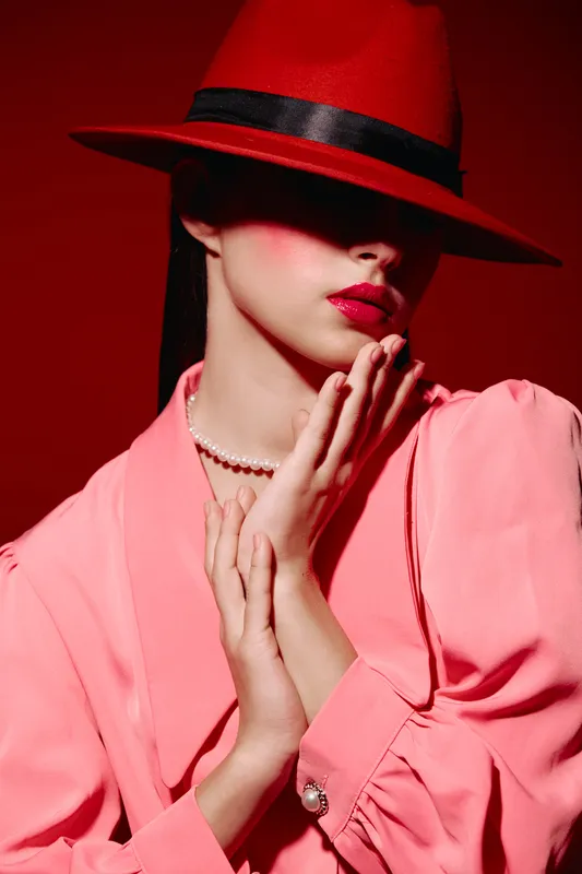 Fashionable woman in pink outfit and red hat partially covers face with shadow. Bold makeup with glossy lipstick and pearl necklace. Concept of beauty, fashion, creativity, style.