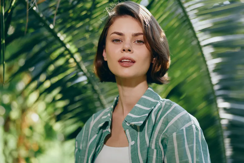 Portrait of a woman with short dark hair wearing a green striped shirt, standing outdoors among lush tropical plants and bright natural light.