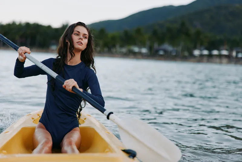 A young woman rows a yellow boat across a calm lake, wearing a dark blue wetsuit with long dark hair, focused expression, steady stroke, ready for adventure.