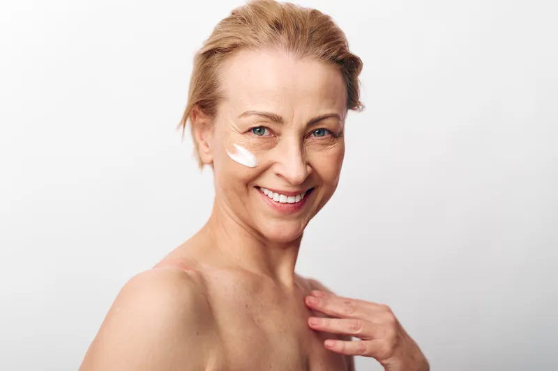 Smiling mature woman with natural skin applies white face cream on cheek. Close-up portrait with bare shoulder on white background. Concept of skincare, beauty routine, hydration, and self-care.