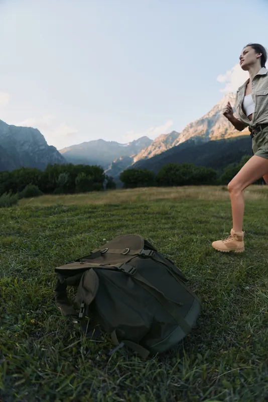 Young woman hiking in mountains stands near backpack on grass field enjoys nature outdoors lifestyle adventure in scenic landscape with clear sky and distant peaks.