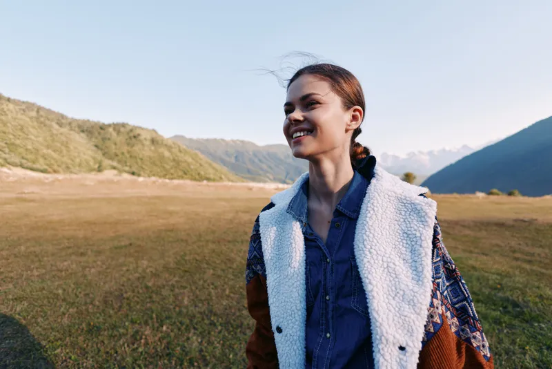 Woman smile portrait in a jacket in a meadow with mountains outdoors, enjoying travel and nature. Young female walking in an open field with scenic hills and clear sky.