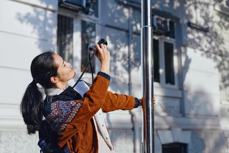 Woman in a warm sweater adjusts equipment on a metal street pole in an urban outdoor setting, near a pale building, illustrating maintenance work and practical workflow.