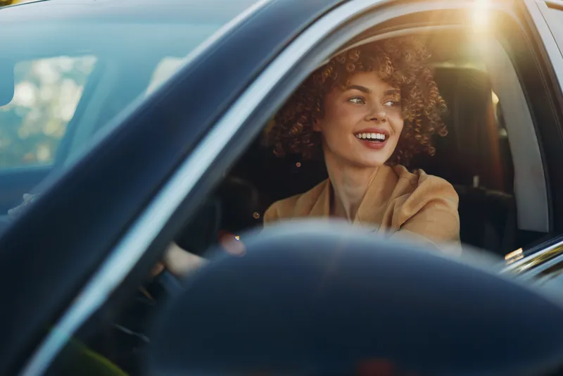 Joyful woman driving a car, sunlit window framing her candid smile, casual outfit, warm retro film tones, outdoor light and gentle breeze, relaxed mood, everyday travel moment captured