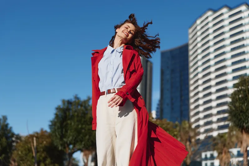 A young woman in a red coat stands confidently in an urban setting, hair swept by wind, embodying bold street style and motion.