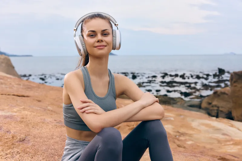 Smiling young woman with headphones enjoying music outdoors, wearing sporty gray outfit, set against a serene beach background with rocks and ocean