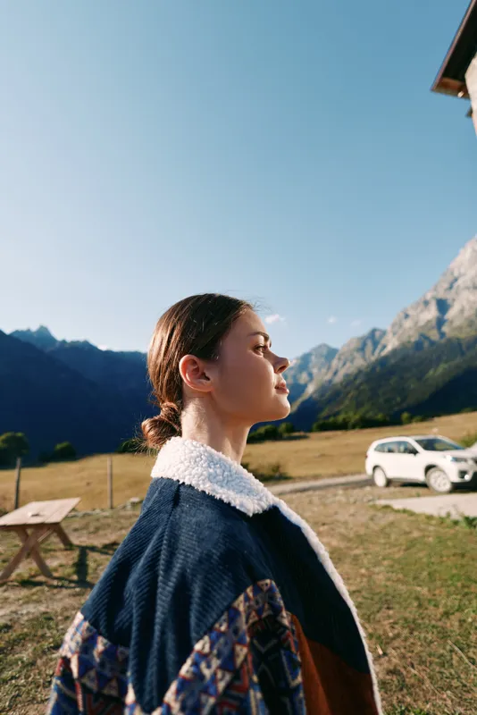 Woman profile in mountains field by a roadside car, wearing a warm jacket and feeling sunlight on her face, outdoor portrait of a young traveler standing near picnic table under clear sky