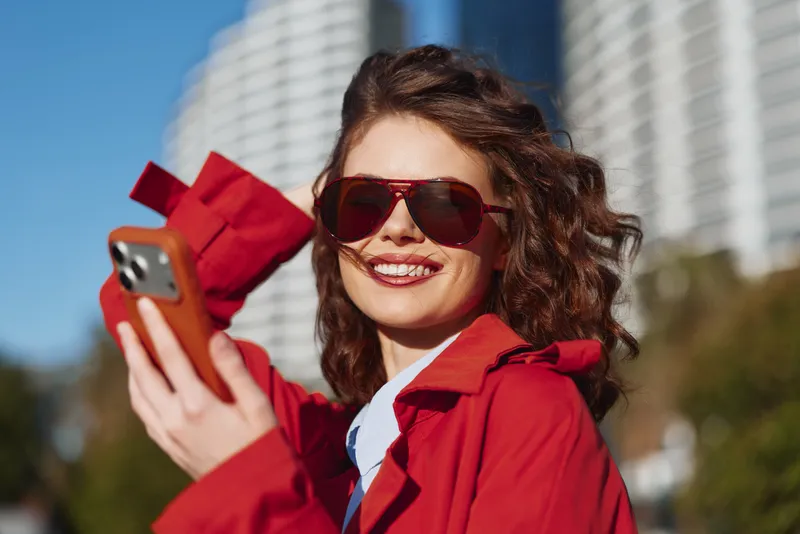 Young woman with curly hair wearing a red coat and sunglasses smiles joyfully while holding a smartphone outdoors on a sunny day in an urban setting.
