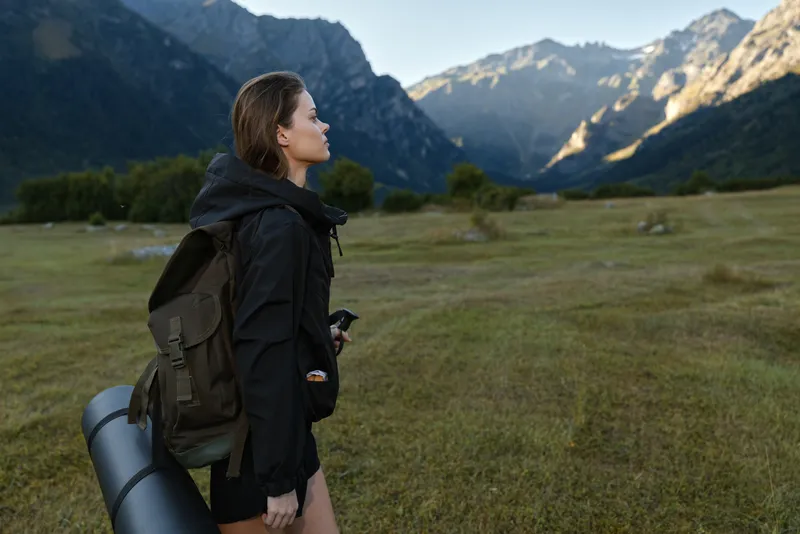 A solitary hiker stands in a wide alpine meadow, backpack secured and a rolled mat at her side, gazing toward distant peaks under clear sky, ready for outdoor adventure.