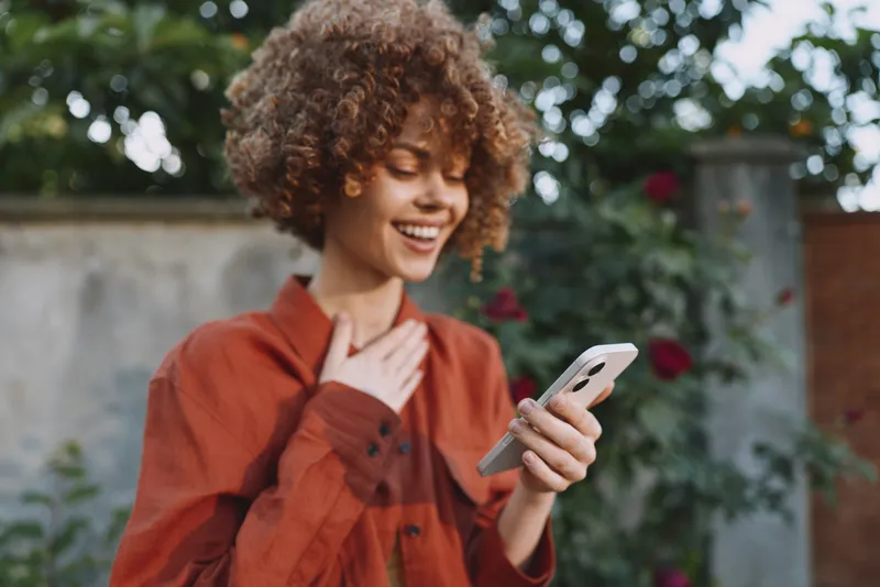 A smiling woman with curly hair checks her smartphone in a sunlit garden, wearing a rust orange shirt. A lifestyle moment highlighting casual joy and digital connection