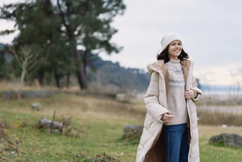 Winter outdoor scene with a smiling woman in a long coat and cozy sweater, standing in a wind swept park by a calm lake, enjoying chilly, serene nature.