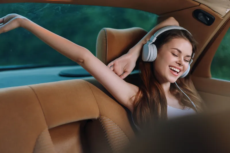 Happy young woman listening to music in a car, enjoying her road trip with headphones, showcasing joy and relaxation in a vibrant green background.