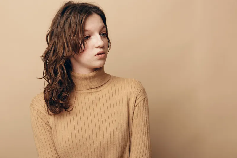 Young woman with wavy brown hair wearing a beige turtleneck sweater looks away thoughtfully against a plain beige background. Concept of contemplation and casual fashion.