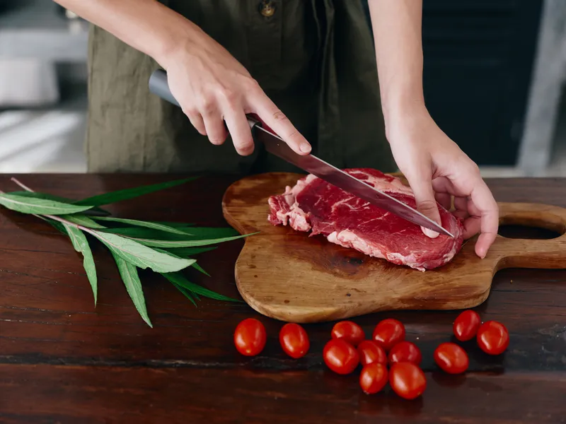 Женщина с ножом в руке нарезает свежее мясо для стейка на жарку на кухне