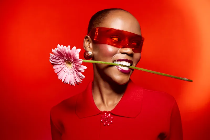 Smiling dark skinned African woman wearing red sunglasses and red shirt holds pink gerbera flower in mouth against red background. Concept of fashion, beauty, and vibrant style.