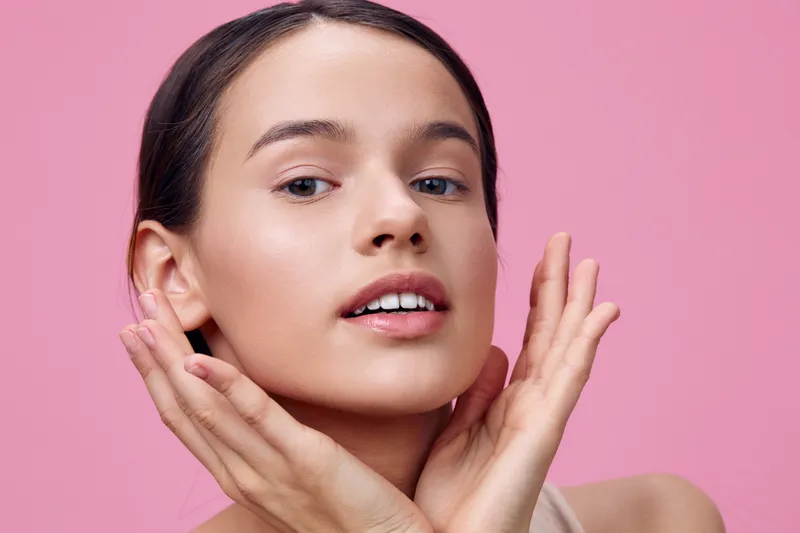 Portrait of a young woman with flawless skin against a soft pink background, showcasing healthy beauty and confidence with a natural makeup look