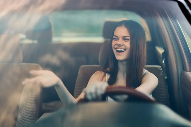 Happy young woman driving a car, enjoying music, with sunlight reflecting on windshield, conveying joy and freedom on a sunny day.