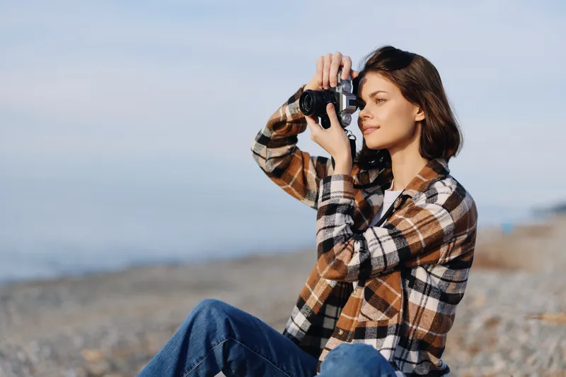 Photographer on a windy beach holds a camera, capturing a relaxed outdoor portrait in a plaid shirt and denim, with calm sea and natural light framing the scene.