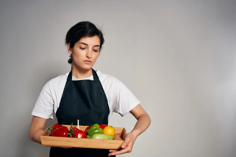 Домохозяйка в черном фартуке на фоне свежих овощей и продуктов