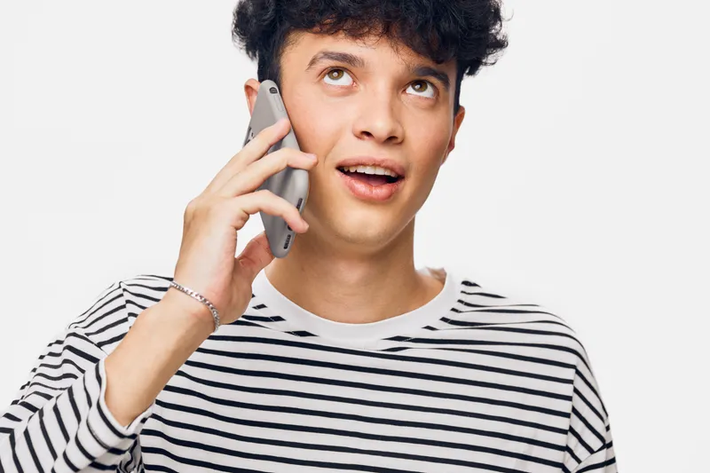 young man talking on smartphone with expressive face, wearing striped shirt, casual style, indoor white background, communication concept