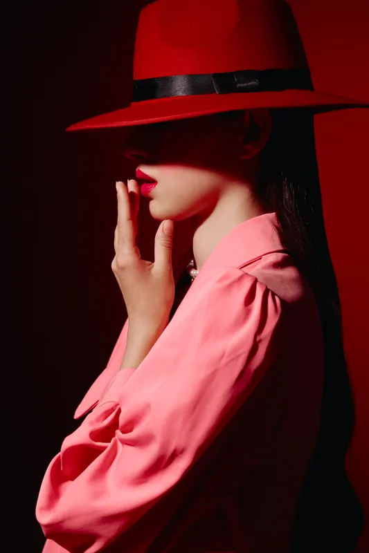 Fashion woman in bright pink blouse and red hat with veil covering eyes touches lips softly. Concept of edgy beauty portrait, creative style, minimalistic red background.