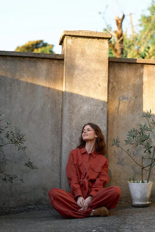 A woman in a red jumpsuit sits cross legged against a weathered wall, relaxed and reflective in an outdoor setting. Concept: everyday woman lifestyle and calm moments
