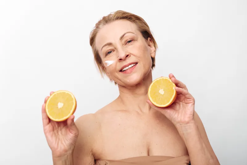Smiling mature woman with skincare cream on cheeks holds two halves of fresh grapefruit. Concept of natural beauty, healthy lifestyle, vitamin c, and facial care for aging skin.